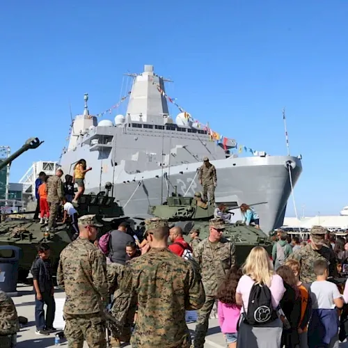 People gather on a dock in front of a large U.S. Navy warship, with soldiers in camouflage and visitors nearby.