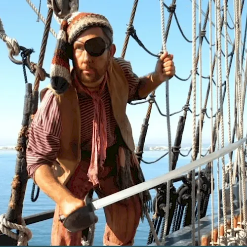 A person in sunglasses and a striped shirt climbs and grips ropes on a sailing ship with the ocean in the background.