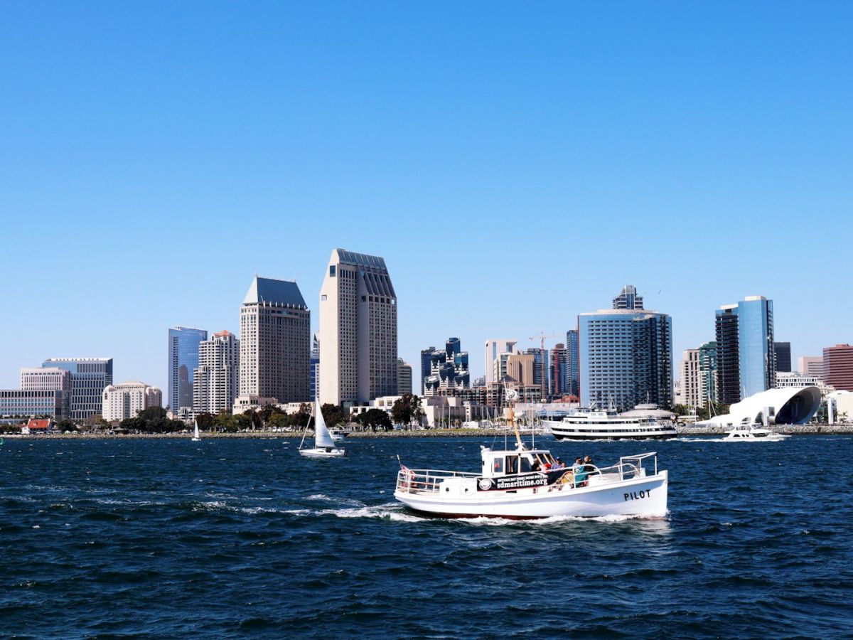 A white research boat labeled PILOT rides across the water before a modern skyline of tall buildings under a clear blue sky.
