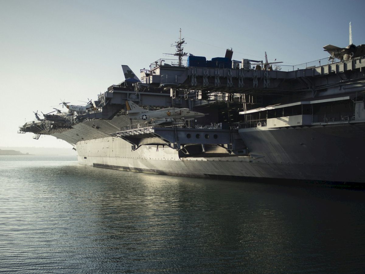A massive aircraft carrier docked, with fighter jets and equipment on its flight deck over calm ocean waters.